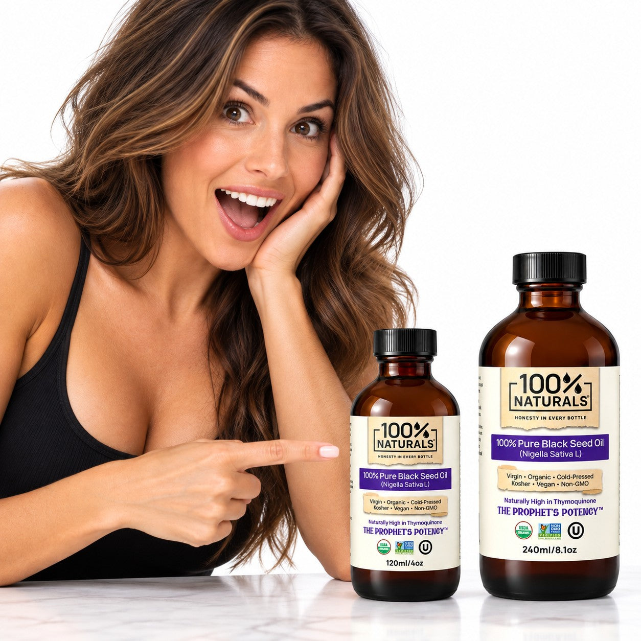 A smiling woman in a black tank top points toward two amber bottles of 100% Naturals seed oils on a white surface against a white background. One bottle is black and the other is amber, both with white labels featuring purple and black text.